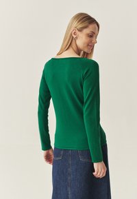 Top verde cu maneci lungi, cu un design ajustat, realizat dintr-un material tricotat moale, având un decolteu tip scoop și detalii cu striații, asortat cu o fustă din denim.