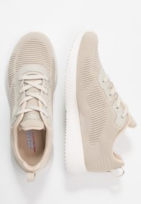 Sneaker in tessuto a maglia beige chiaro con suole bianche, lacci, design testurizzato nella parte superiore e solette in schiuma memory visibili all'interno.