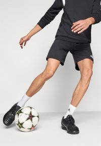 Μαύρα αθλητικά παπούτσια, γκρι σόρτς και μαύρο φούτερ με λευκά σημεία· μια μπάλα ποδοσφαίρου στο έδαφος, που φέρει ένα αστέρι σχέδιο.