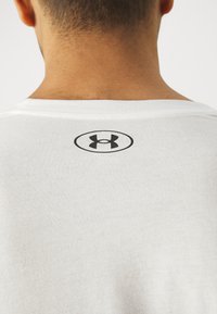 Primer plano de la espalda de una persona que lleva una camiseta blanca con un logo negro de Under Armour debajo del escote.