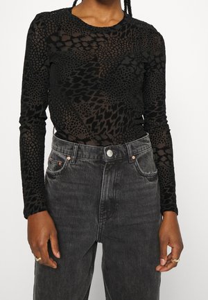 Personne portant un haut noir transparent à manches longues avec un motif animalier et un jean noir délavé taille haute, les mains reposant sur les côtés.