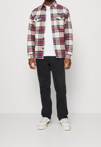 Beżowa koszula z flaneli w kratę w odcieniach czerwieni i niebieskiego; biała koszulka; czarne spodnie; jasne sneakersy. Koszula zapinana na guziki z dwiema kieszeniami na piersiach.