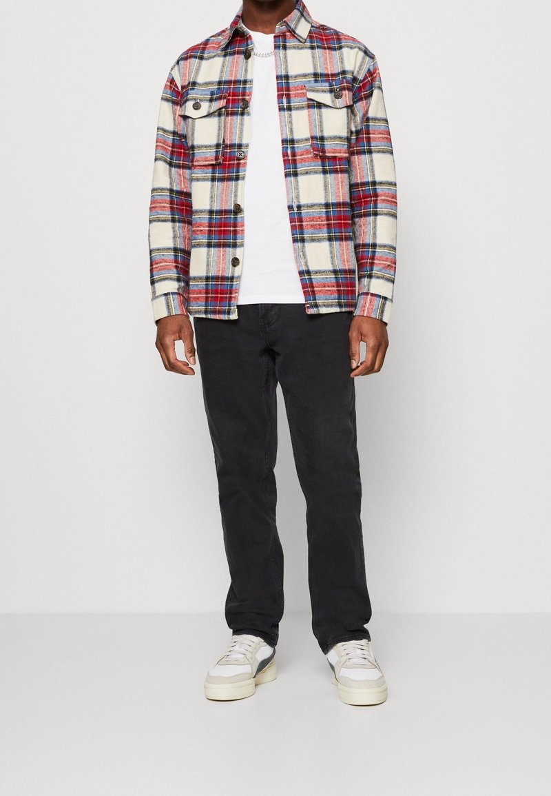 Beżowa koszula z flaneli w kratę w odcieniach czerwieni i niebieskiego; biała koszulka; czarne spodnie; jasne sneakersy. Koszula zapinana na guziki z dwiema kieszeniami na piersiach.