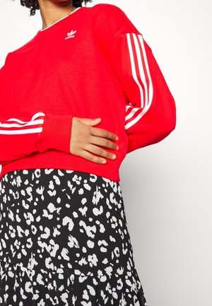 Person wearing a red Adidas sweatshirt with white stripes on sleeves and a black and white spotted skirt, hand resting on the waist.