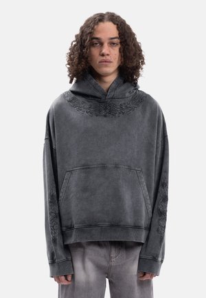 Joven con cabello rizado que lleva una sudadera con capucha oversize de color gris oscuro con diseños tribales sutiles en el pecho y las mangas, y pantalones grises descoloridos.