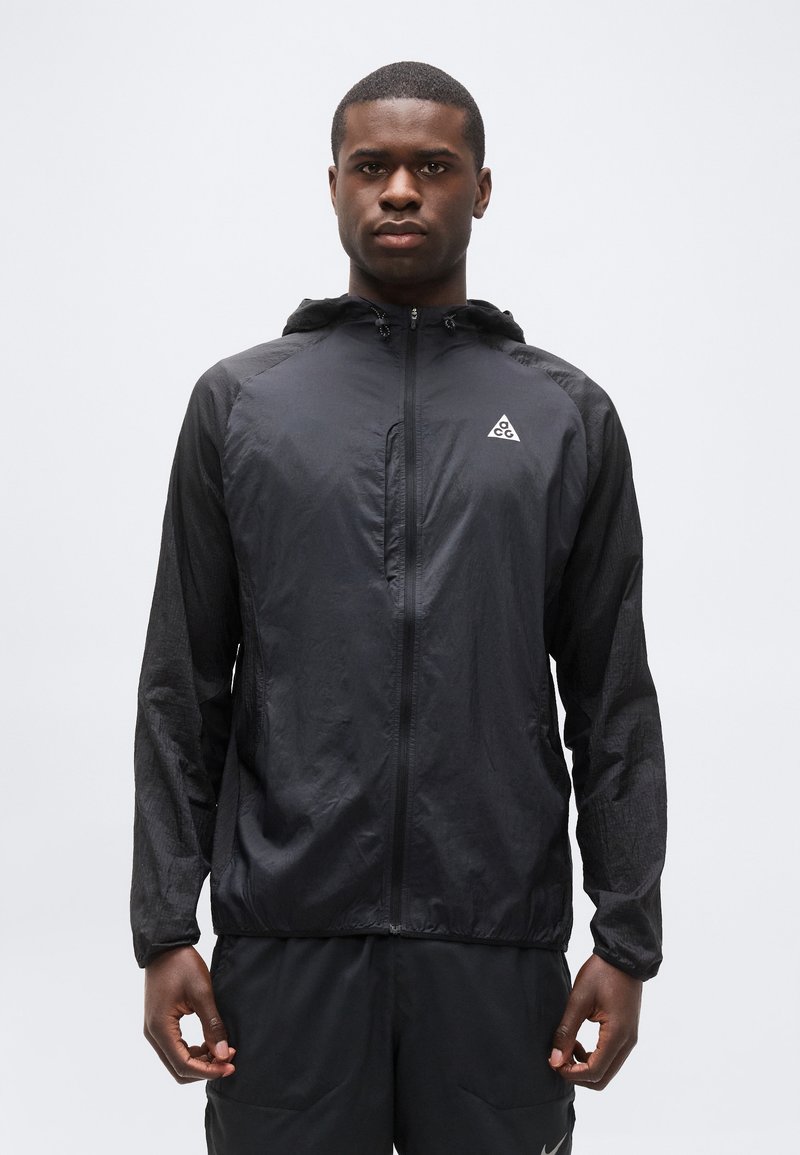 Άνδρας που φοράει μαύρο ελαφρύ μπουφάν με κουκούλα, φερμουάρ μπροστά και λογότυπο ACG στο στήθος, στέκεται μπροστά σε απλό φωτεινό φόντο.