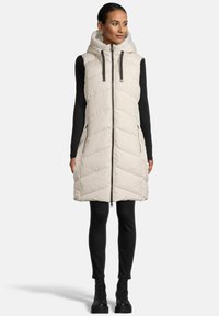 Femme portant un gilet sans manches crème rembourré avec capuche, un haut à manches longues noir à côtes, un pantalon noir et des bottes noires à lacets, se tenant droite.
