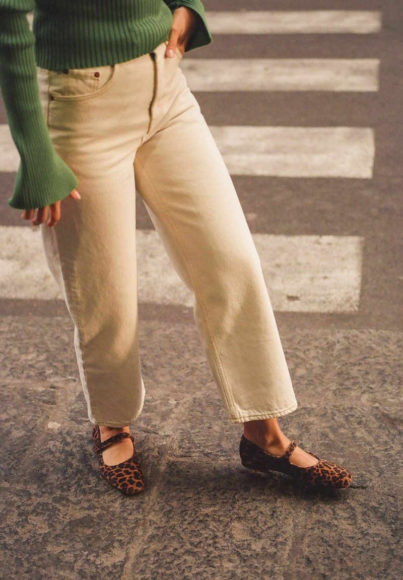 Personne portant un pantalon beige, un pull côtelé vert et des chaussures Mary Jane à imprimé léopard, debout dans une rue avec un passage piéton en arrière-plan.