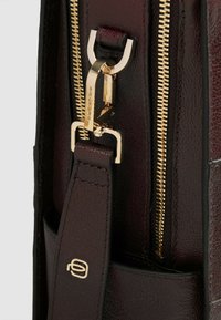 Bordeaux synthetische leren tas met detail van een goudkleurige rits, klembevestiging en een gladde textuur; voorzien van een merkaccent op de riem.