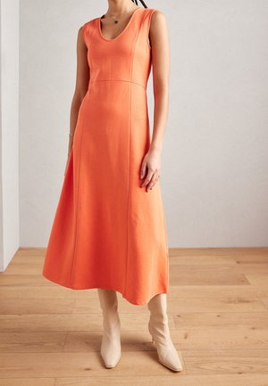 Vrouw draagt een mouwloze oranje midi-jurk met een ronde halslijn en beige laarzen met hakken, staand op een houten vloer tegen een lichte muur.