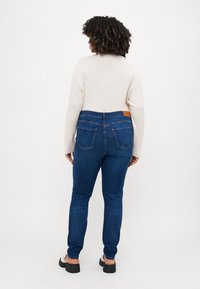 Dark blue denim jeans with a slim fit, featuring a mid-rise waist, back pockets, and a brown leather tag at the waistband. Textured light sweater.