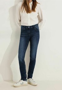 Mörkblå denimjeans med fem fickor, lätt blekning och en figurnära passform. Matchas med en vit skjorta med knappar och vita sneakers.