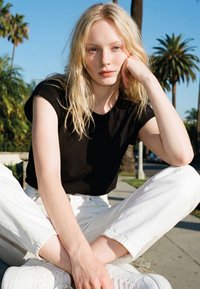 Plavuša u crnoj majici i bijelim hlačama sjedi prekriženih nogu na otvorenom, na sunčan dan, s palmama u pozadini.