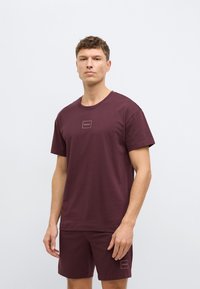 Μπορντό βαμβακερό μπλουζάκι με ριμπ λαιμόκοψη και μικρό επάνω λογότυπο στο στήθος, σε συνδυασμό με σορτς στο ίδιο χρώμα, και τα δύο μονόχρωμα.