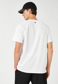 Camiseta atlética blanca de manga corta con mangas raglán y dobladillo redondeado. Presenta un pequeño logo en la parte posterior cerca del escote.