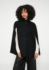 Μαύρη μάλλινη κάπα με ψηλό γιακά και δύο κουμπιά στο μπροστινό μέρος, με ανοιχτές πλευρές, φορεμένη πάνω από ένα κρεμ πουλόβερ και συνδυασμένη με μαύρο παντελόνι.