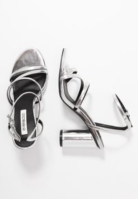 Srebrne sandały na wysokim obcasie z błyszczącym wykończeniem, z wieloma wąskimi paskami i masywnym metalicznym obcasem. Czarne wkładki.