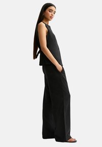 Zwarte mouwloze top met een getailleerd ontwerp en een zijstrikdetail, gecombineerd met zwarte brede broek en minimalistische sandalen.