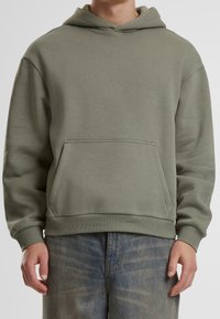 Felpa con cappuccio verde oliva con tasca frontale, polsini e orlo a coste. Realizzata in tessuto morbido e strutturato con una vestibilità comoda.