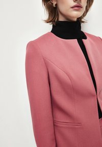 Giacca tailored rosa con spalle strutturate, tessuto testurizzato e scollatura arrotondata. Presenta un dolcevita nero sotto e due tasche frontali.