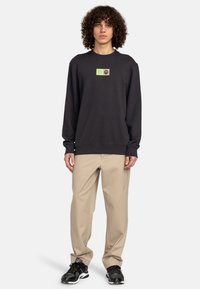 Black sweatshirt with a large green and beige graphic, crew neck, ribbed cuffs, paired with beige trousers and black athletic shoes.