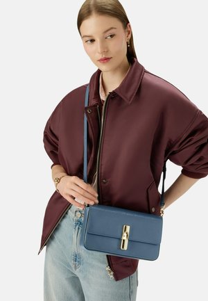 Sac bandoulière en cuir bleu avec une texture lisse, de forme rectangulaire et avec des accessoires en doré. Porté avec une veste bordeaux et un denim clair.