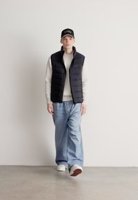 Melns polsterēts veste pār krēmveida ribotu džemperi, kopā ar gaiši zilām vaļīgām džinsām un baltām sporta kurpēm. Melna cepure ar baltiem akcentiem.