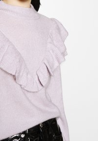 Pull en tricot violet clair avec des détails volantés le long des épaules et de la poitrine, associé à un pantalon noir brillant.