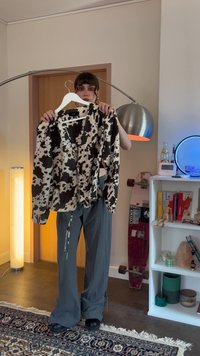 Persoon die een zwart-wit koeienprint blouse vasthoudt aan een hanger in een moderne kamer met een skateboard, lamp en boekenkast op de achtergrond.