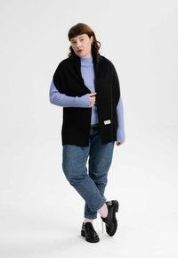 Zwarte gebreide sjaal gedragen over een lichtblauwe geribde trui, gecombineerd met blauwe jeans en zwarte veterschoenen. De sjaal heeft een label.