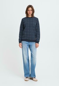 Un sweat bleu marine avec des motifs en forme d'étoile, un col rond et des poignets côtelés, associé à un jean droit bleu délavé et des chaussures beiges.