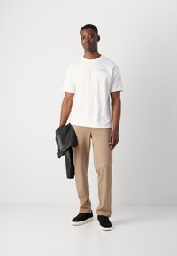 Hvit t-skjorte med korte ermer og subtil logo, beige skreddersydde bukser, svart skinnjakke over den ene armen, og svarte joggesko med hvite såler.