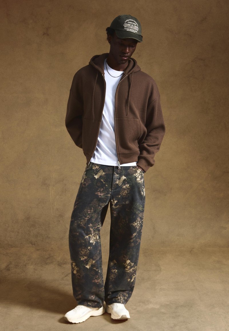Brown ribbed zip-up hoodie over a white T-shirt, paired with loose-fitting dark denim pants featuring a multi-colored camo pattern. White sneakers.