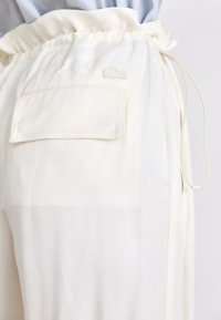 Kremfargede joggebukser med elastisk liv, sidelommer og en baklomme med et preget logo. Glatt, lett stoff.