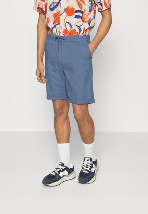 Hellblaue Shorts aus einem weichen Stoff, mit einem elastischen Bund und einem Kordelzug. Dazu weiße Socken und marineblaue Sneakers.