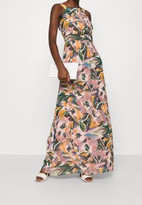 Vestido largo sin mangas con diseño de flores en tonos rosa, naranja y verde, confeccionado con tela ligera. Bolso de mano blanco, combinado con sandalias blancas.