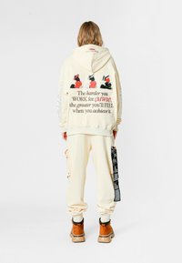 Persona con sudadera y pantalones color beige con gráficos en rojo y negro y texto motivacional en la parte de atrás, usando botas marrones, de pie sobre fondo blanco.