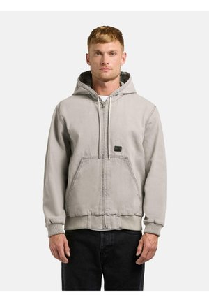 Ung mand iført en lysegrå lynlåshættetrøje med forlommer og snørejustering i hætten, kombineret med sorte bukser, set forfra.