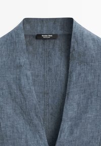 Límec a výstřih tmavě šedého lněného saka ukazující vnitřní štítek s názvem značky "Massimo Dutti" a textem "Vyrobeno v Číně".