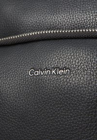 Primo piano di pelle nera texturizzata con una cerniera argentata e il logo "Calvin Klein" ben in vista in bianco.
