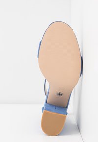 Sandale à talon en suede bleu avec une semelle en caoutchouc beige lisse, présentant un talon rectangulaire et un design minimaliste, montée sur un fond blanc.