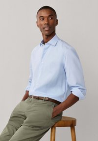 Mann trägt hellblaues Hemd und olivgrüne Hose, sitzt auf einem hölzernen Hocker, Hände in den Taschen, schaut seitlich vor einfachem Hintergrund.