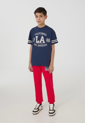 Maglietta navy con grafica bianca che riporta la scritta "California LA 86", abbinata a pantaloni della tuta rossi con logo bianco. Sneakers in bianco e nero.
