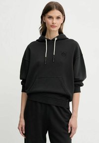 Donna che indossa una felpa nera con cordini bianchi e pantaloni neri coordinati, in piedi davanti a uno sfondo chiaro e uniforme.