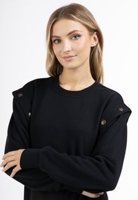 Zwarte sweatshirt met ronde halslijn, schouderdetails met gouden knopen, en lange mouwen met een strakke manchette. Zachte textuur is duidelijk zichtbaar.