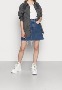 Frau trägt ein graues Hemd mit Knöpfen, ein weißes Batik-Bauchfreies Top, einen ausgefransten Jeansrock und weiße Plateau-Stiefel, steht vor einem schlichten Hintergrund.