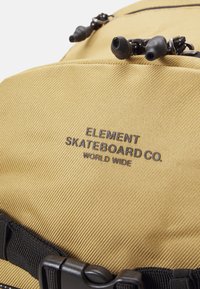 Žuti ruksak od tkanine s crnim patentnim zatvaračima i remenima. Ima logo "ELEMENT SKATEBOARD CO." u tamnom printu, teksturirana površina.