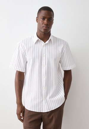 Homme debout contre un mur blanc, portant une chemise blanche à manches courtes avec de fines rayures verticales et un pantalon marron.