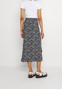 Mujer de pie de espaldas, vistiendo una blusa blanca de manga corta, una falda midi negra con estampado floral blanco y sandalias blancas con hebilla.