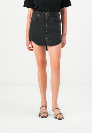 Kvinne som har på seg en svart knappefrosset denim mini skjørt og svarte stroppede hælsandaler mot en enkel hvit bakgrunn.